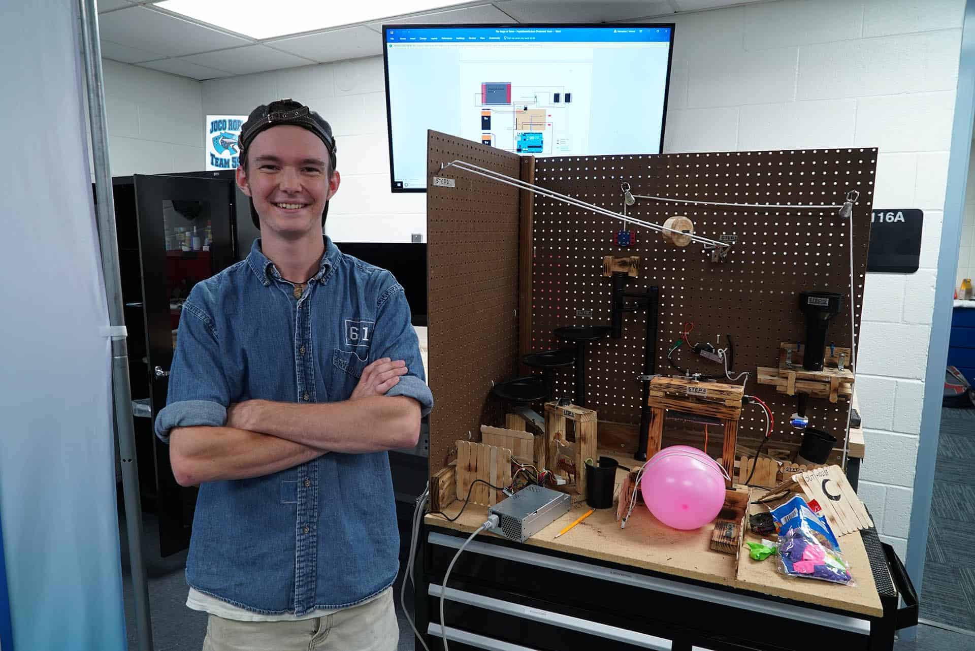 a student stands arms folded in front of Rube Goldberg machine