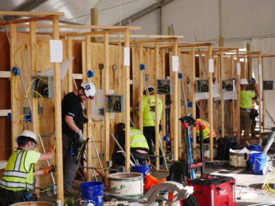 Apprentices wearing hard hats and bright colors work in wooden stalls during an electrical competition. Some are standing while others are crouching.