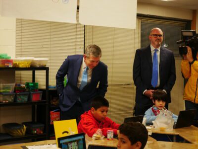 Gov. Roy Cooper leans over a table to watch two third graders work on computers as they eat their breakfasts.