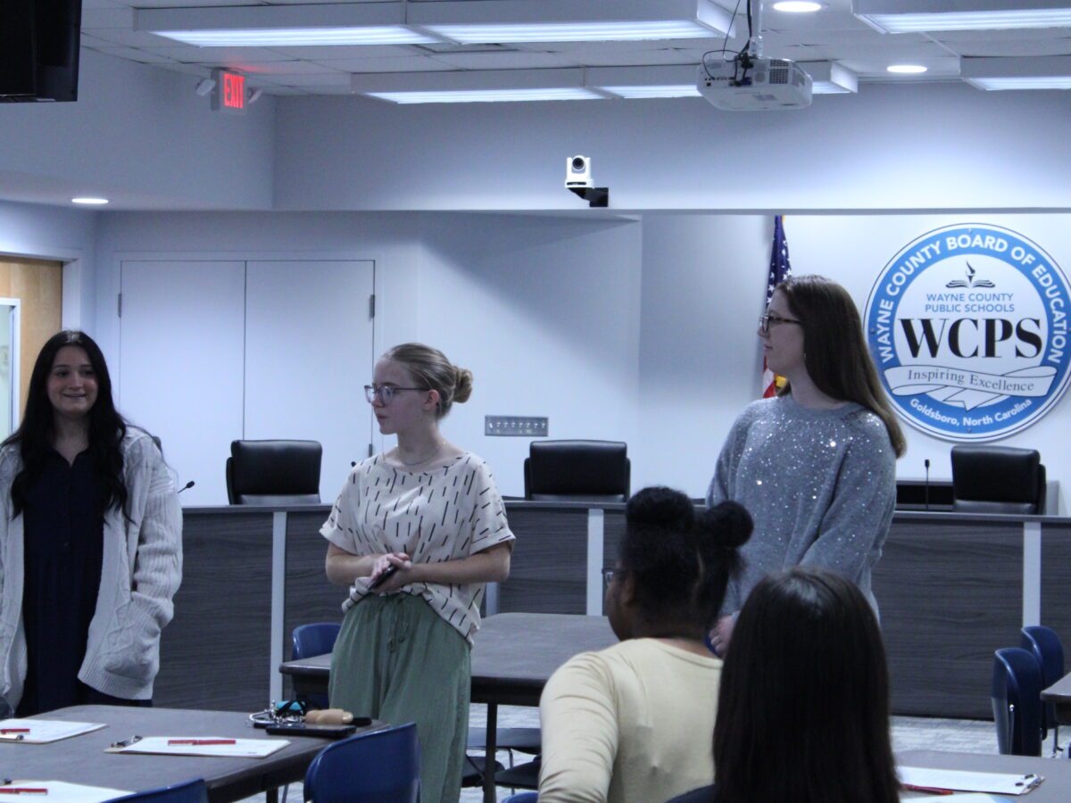 three women standing in front of Wayne County Board of Education seal and a desk with four swivel chairs in a board room.