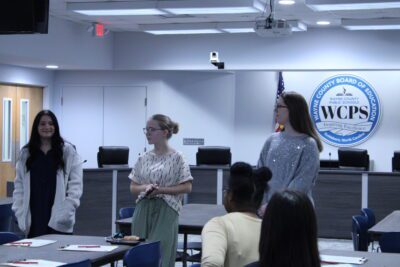 three women standing in front of Wayne County Board of Education seal and a desk with four swivel chairs in a board room.