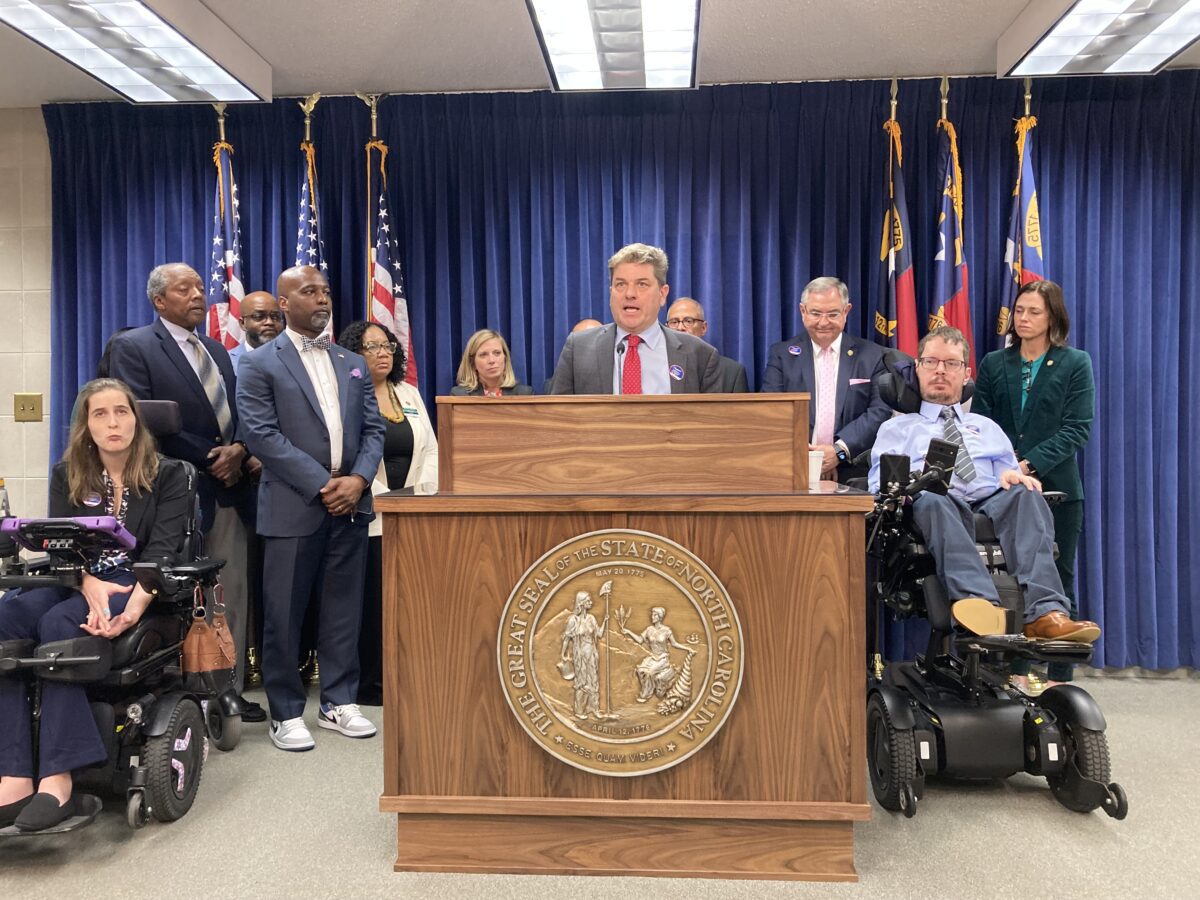man standing at podium surrounded by half dozen people and state flags