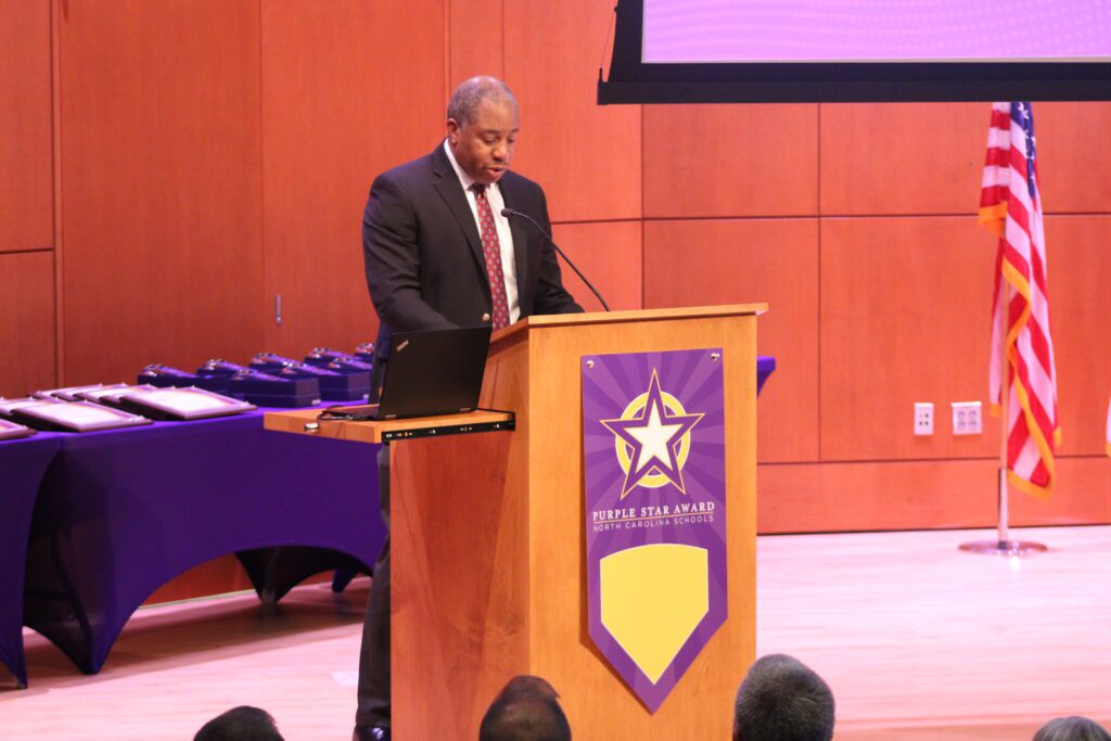 man standing at podium in front of purple table and american flag