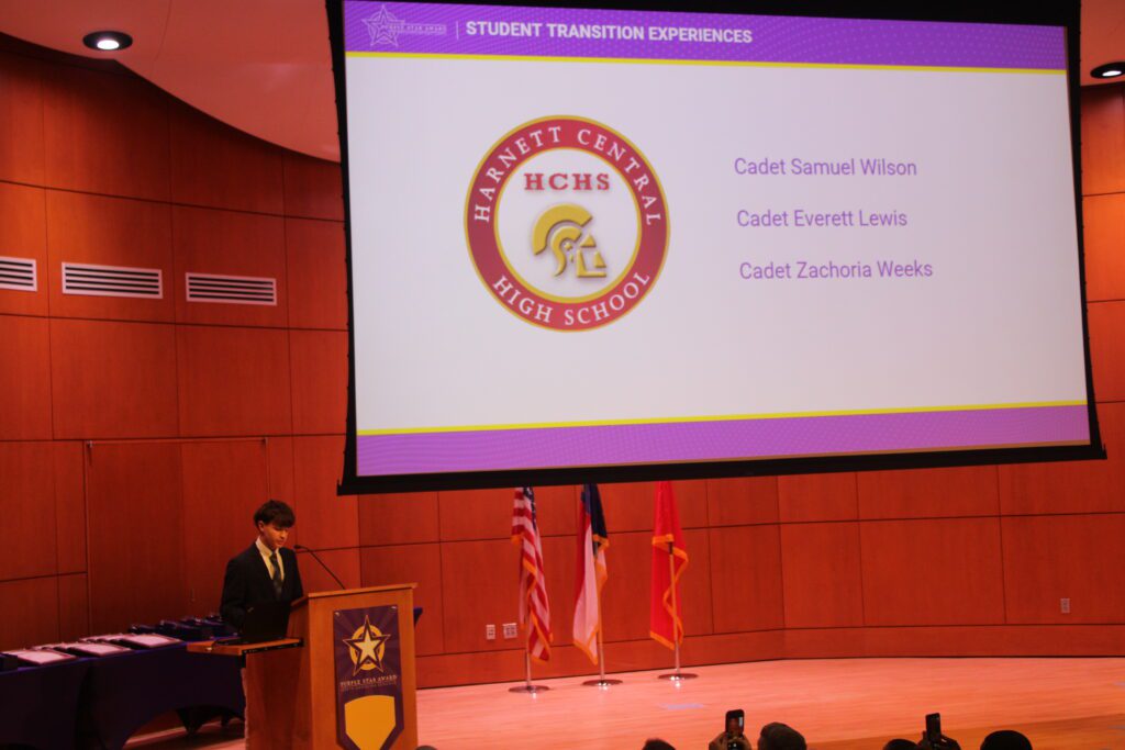 student standing on stage under purple slide show with his name and high school emblem