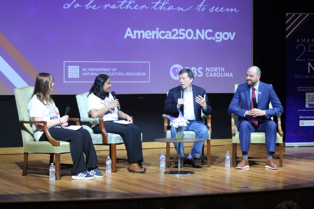 four people sit on stage in front of pbs north Carolina logo with mics in their hands. Burns is using his hands to explain his point of view.