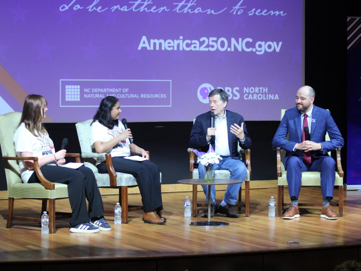 four people sit on stage in front of pbs north Carolina logo with mics in their hands. Burns is using his hands to explain his point of view.