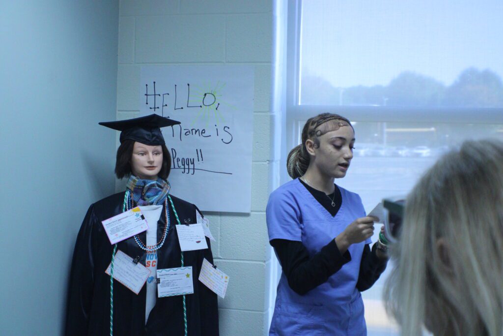 student in scrubs speaking in front of mannequin with notecards pinned to it