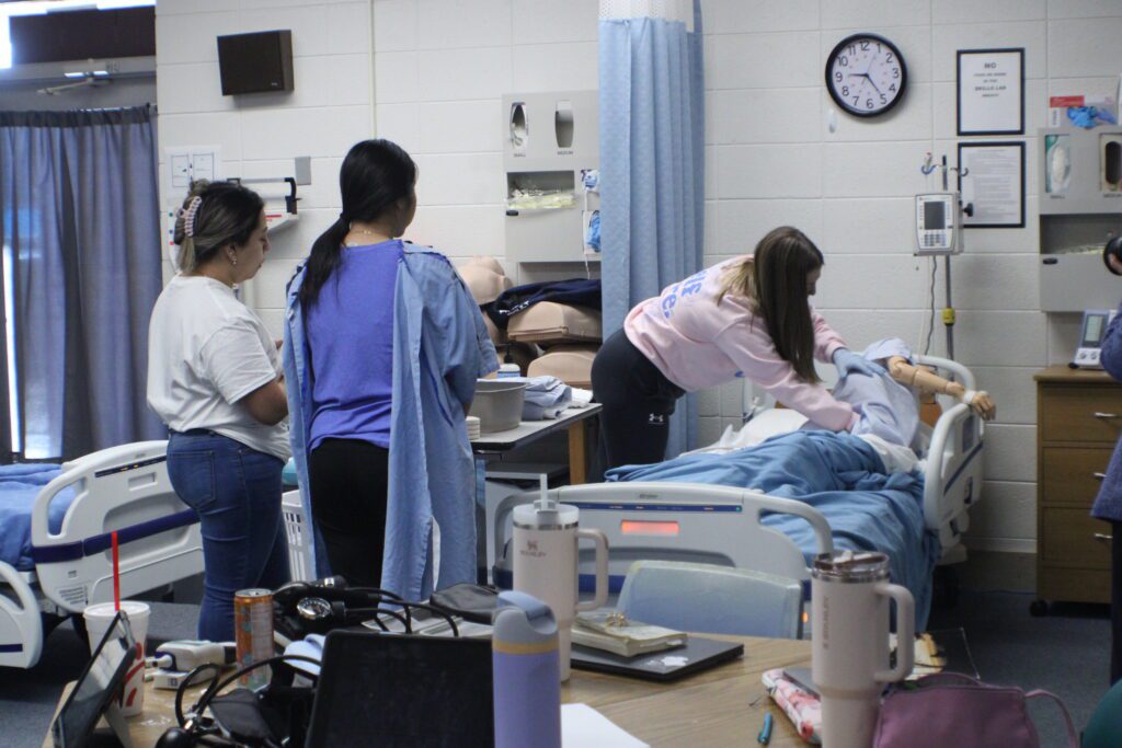 student inspecting a mannequin in a hospital bed while two other students watch