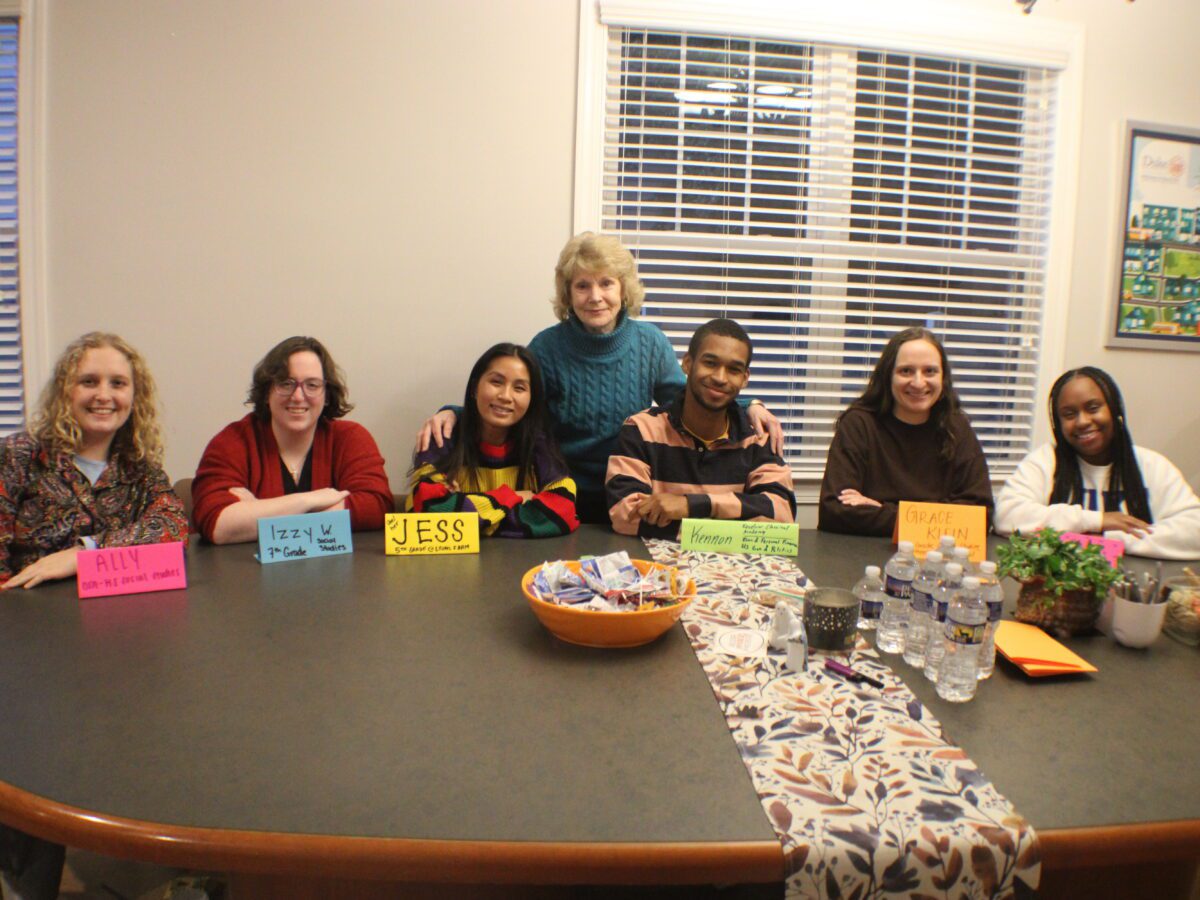 seven people sit and stand around an oval table with name tags
