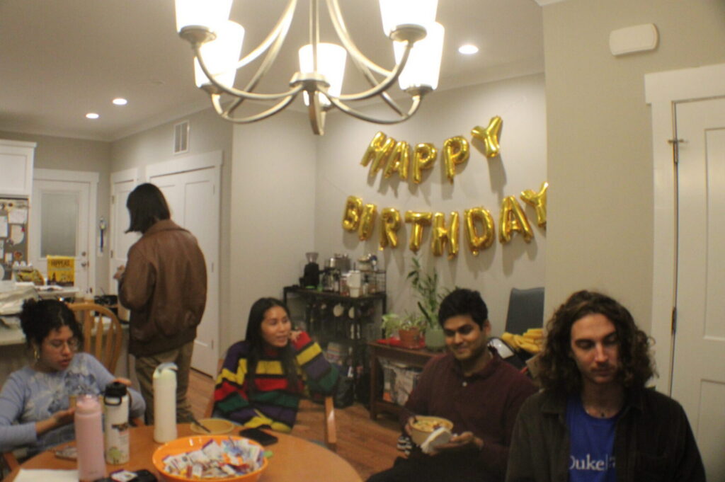 people sitting at a round table with happy birthday balloons taped to a wall.