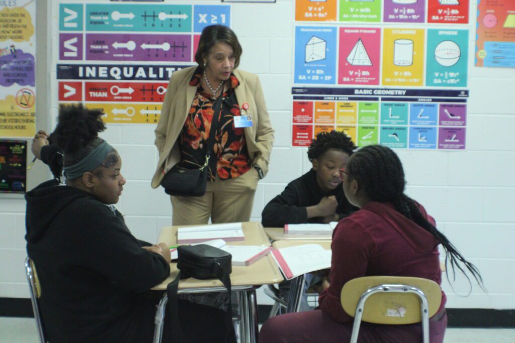 woman standing at desk table with four students doing math