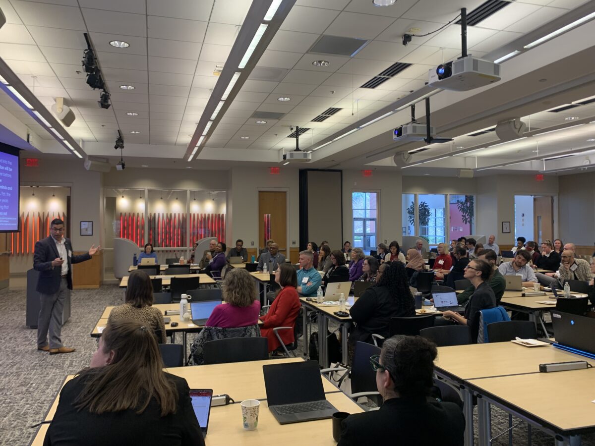 Man standing in center of room of people sitting at desks