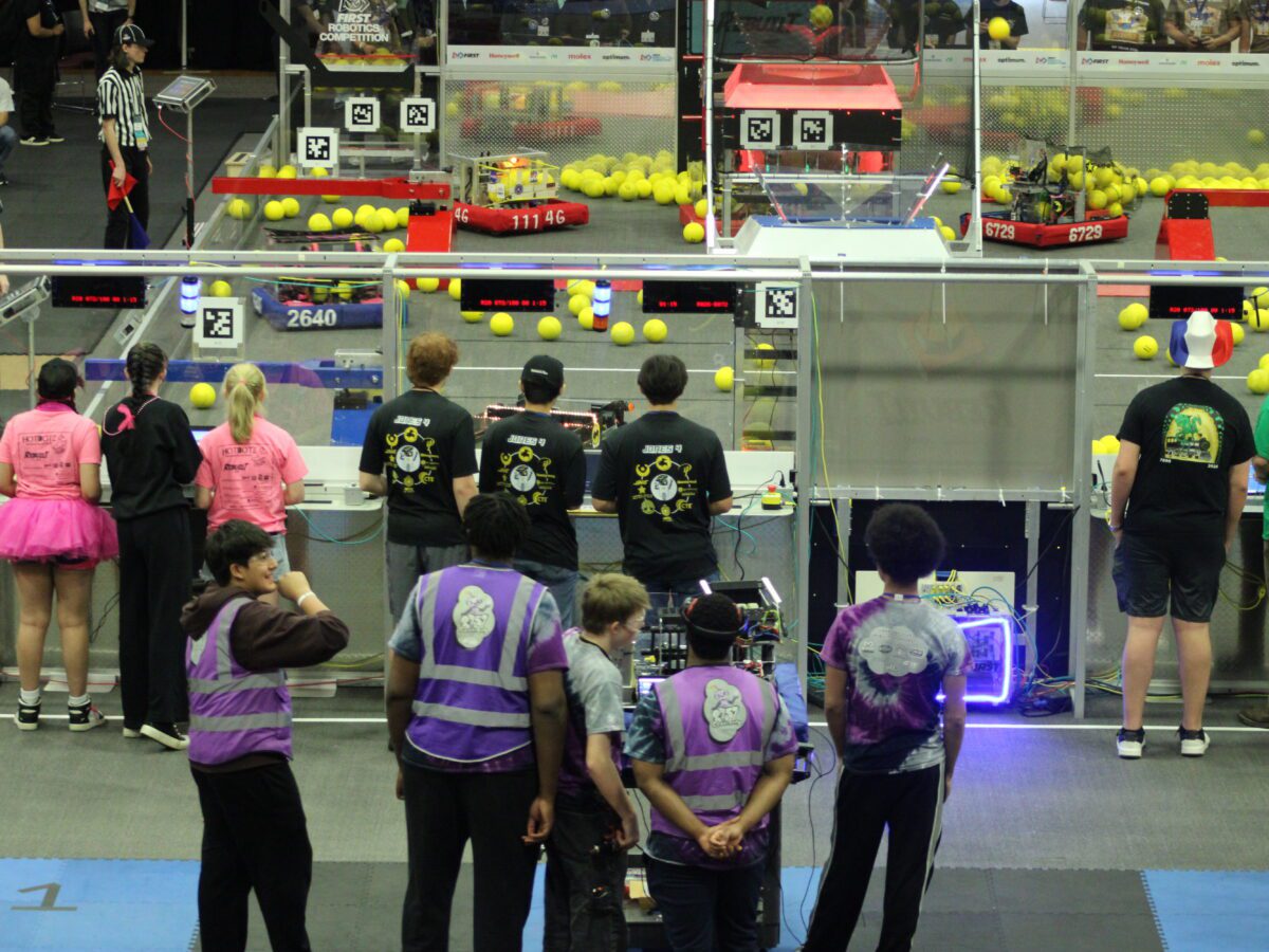 students standing behind the glass of an arena filled with large tennis balls and square robots with wheels