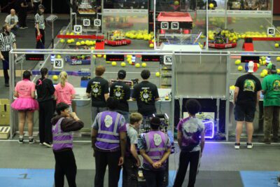 students standing behind the glass of an arena filled with large tennis balls and square robots with wheels
