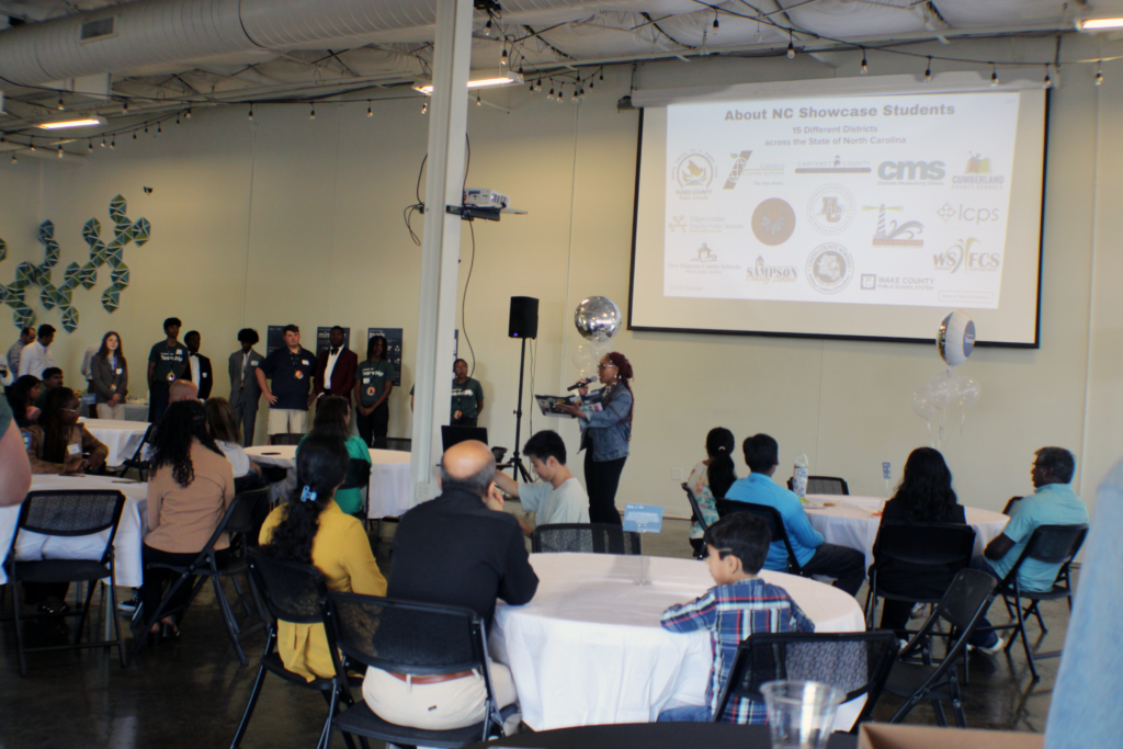 woman standing in front of room with roundtables and projector screen with people standing on wall