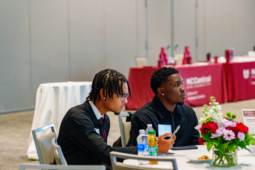 Two individuals sit at a table during an event, one looking down at a phone while the other gazes ahead, with a floral arrangement and branded tablecloth nearby.